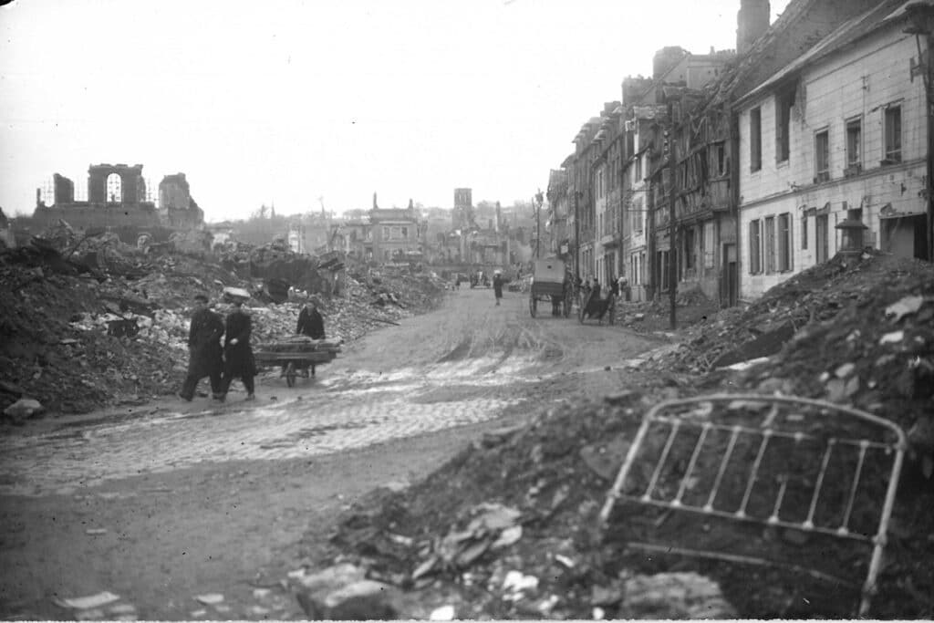 Route de Caen après les bombardements de juin 1944. Au fond à gauche ruines de l'église Saint-Désir. Au bout de la rue couvent des Bénédictines. On aperçoit des personnes avec des brouettes ou des charettes. Une armature de lit au premier plan. .Don à la médiathèque par M. Pierre Lebaud